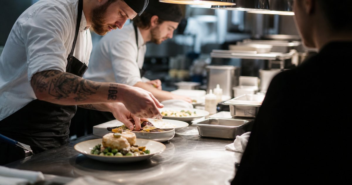 Line cooks plating dishes at a stainless steel pass while a server waits on the other side, heat lamps glowing, busy restaurant kitchen