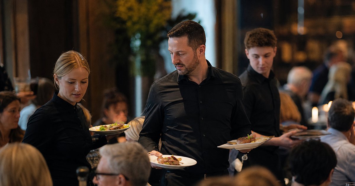 Restaurant floor manager in a dark shirt carrying plates to a table alongside servers during a busy dinner service, dining room full of guests