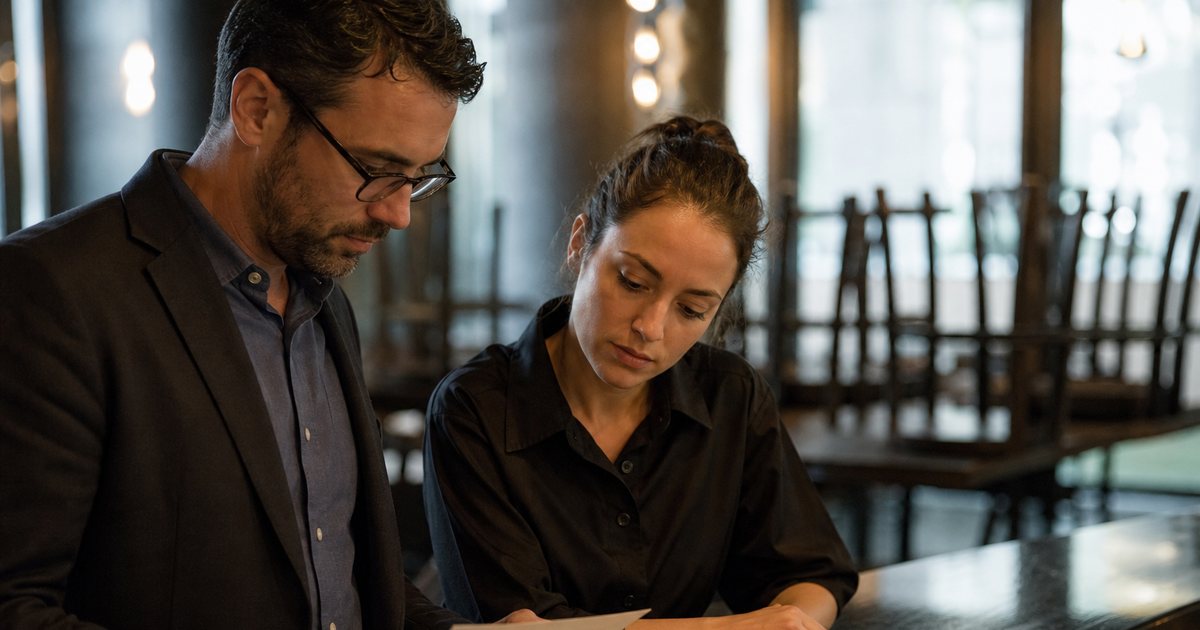 Restaurant manager and a server having a calm conversation at a bar counter after closing, two glasses of water between them, chairs stacked on tables in the background