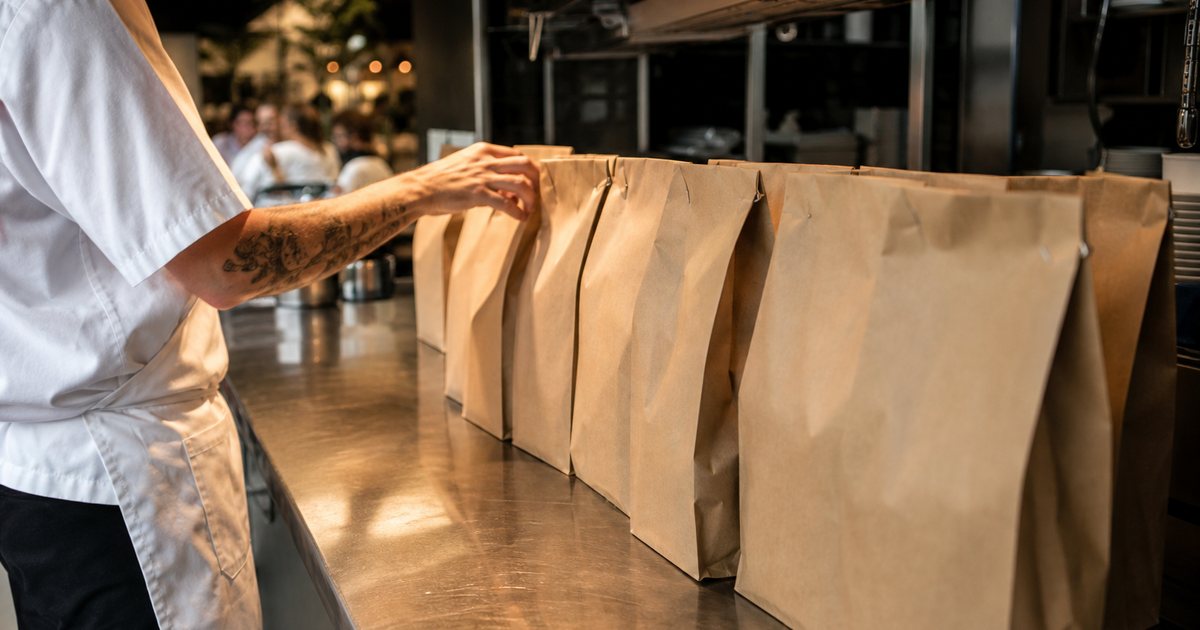 Plain brown paper takeout bags stapled shut and lined up along a stainless steel kitchen pass, a cook in a white apron reaching for one, warm overhead heat lamps, blurred dining room visible in the distance